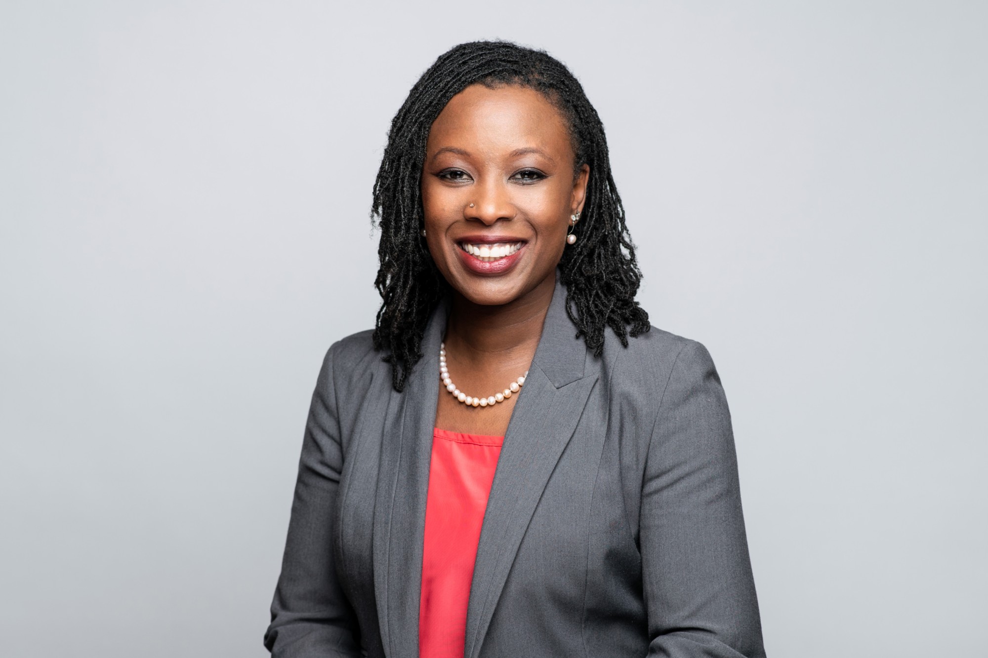 Yabome Gilpin-Jackson is pictured seated and smiling, wearing a grey blazer and red blouse against a light grey backdrop